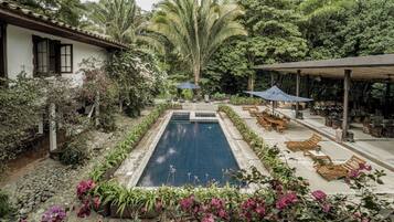 Ein Pool, umgeben von üppiger Vegetation, Holzliegen und einer überdachten Terrasse.