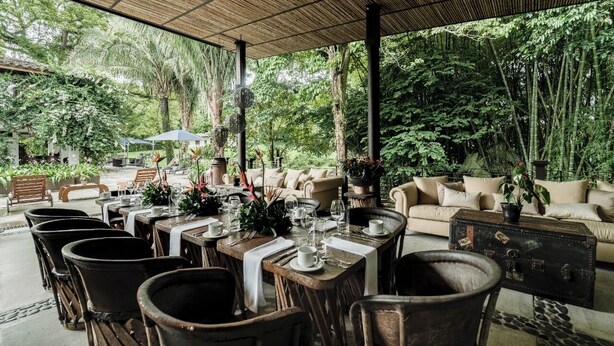Een buitenontbijtplaats met tafels voor twee personen, omringd door stoelen en banken, met uitzicht op weelderige begroeiing.