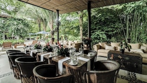 Een buitenontbijtplaats met tafels voor twee personen, omringd door stoelen en banken, met uitzicht op weelderige begroeiing.