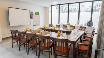 Sala de conferência com quadro branco, amplas janelas com vista para a cidade, mesas e cadeiras de madeira e uma máquina de café.