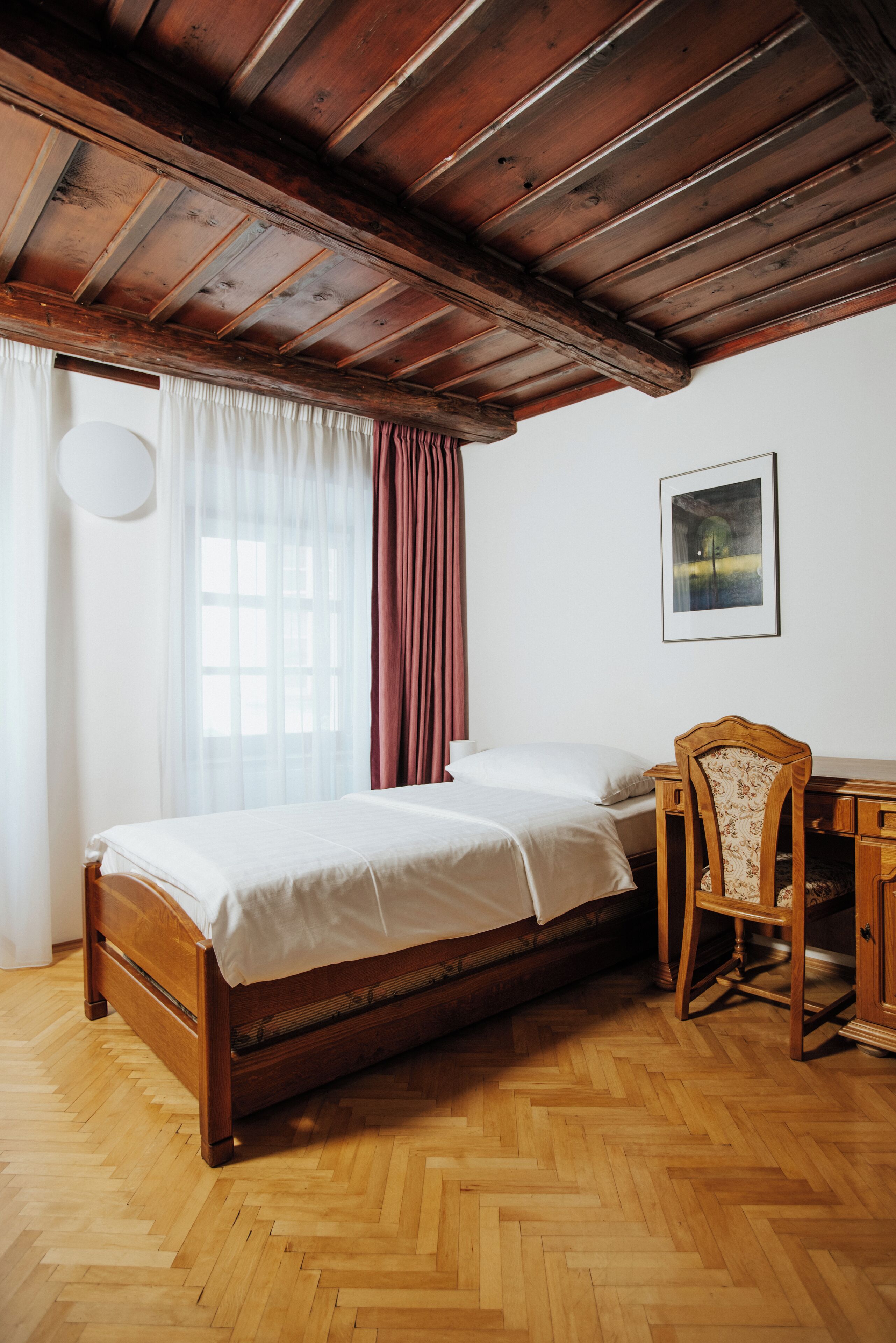 A bedroom with a wooden bed, a chair, a desk, and a window with curtains.