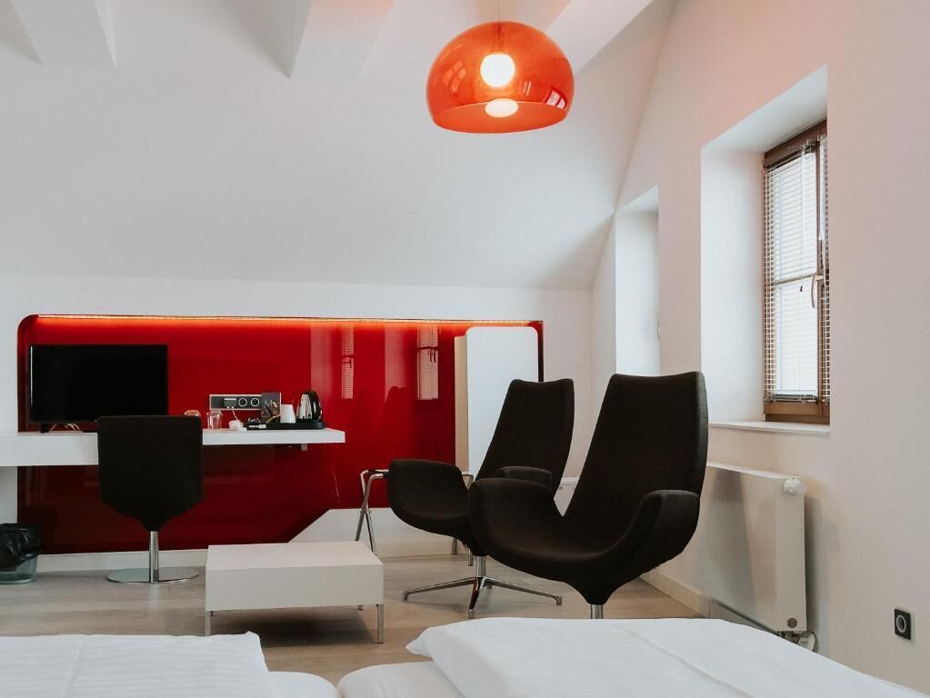 Una habitación de hotel moderna con una pared roja de acento, un televisor de pantalla plana, una silla negra, un sofá blanco y un ventanal grande.