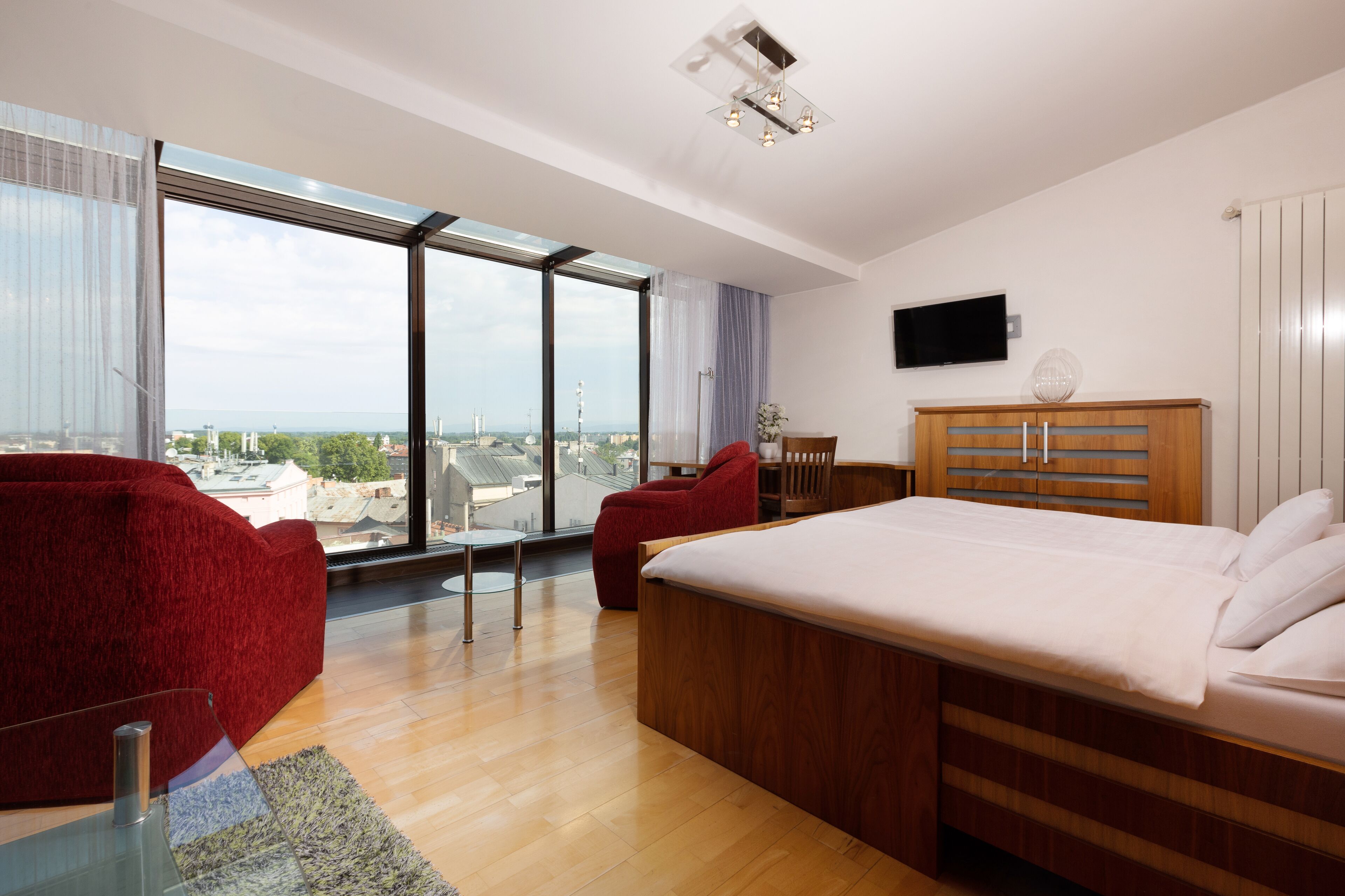 A modern bedroom with a large bed, a red chair, and a view of the city through large windows.