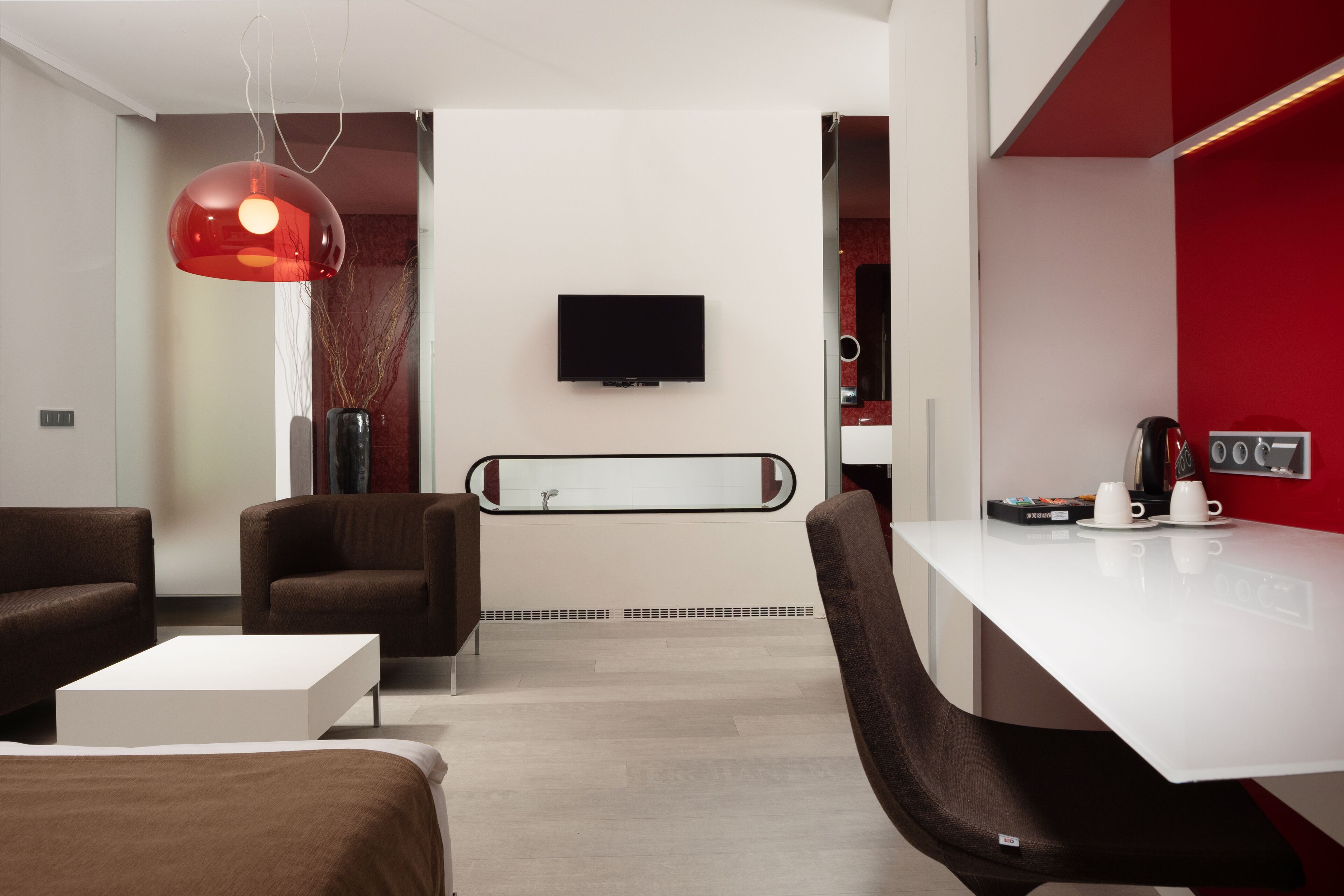 Quarto de hotel moderno com cama branca, sofás castanhos, mesa de centro branca, luminária pendente vermelha, televisão de tela plana e uma parede de destaque vermelha.