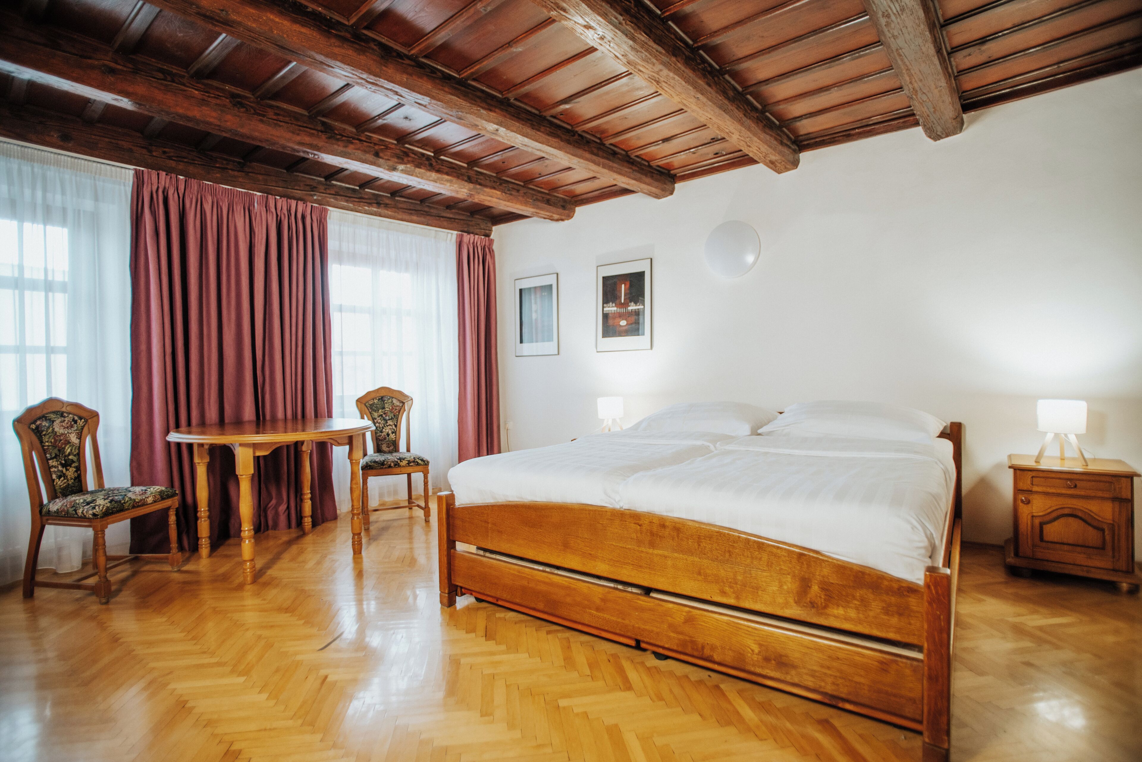Quarto com cama de madeira, mesa de madeira, duas cadeiras e mesa de cabeceira com abajur.