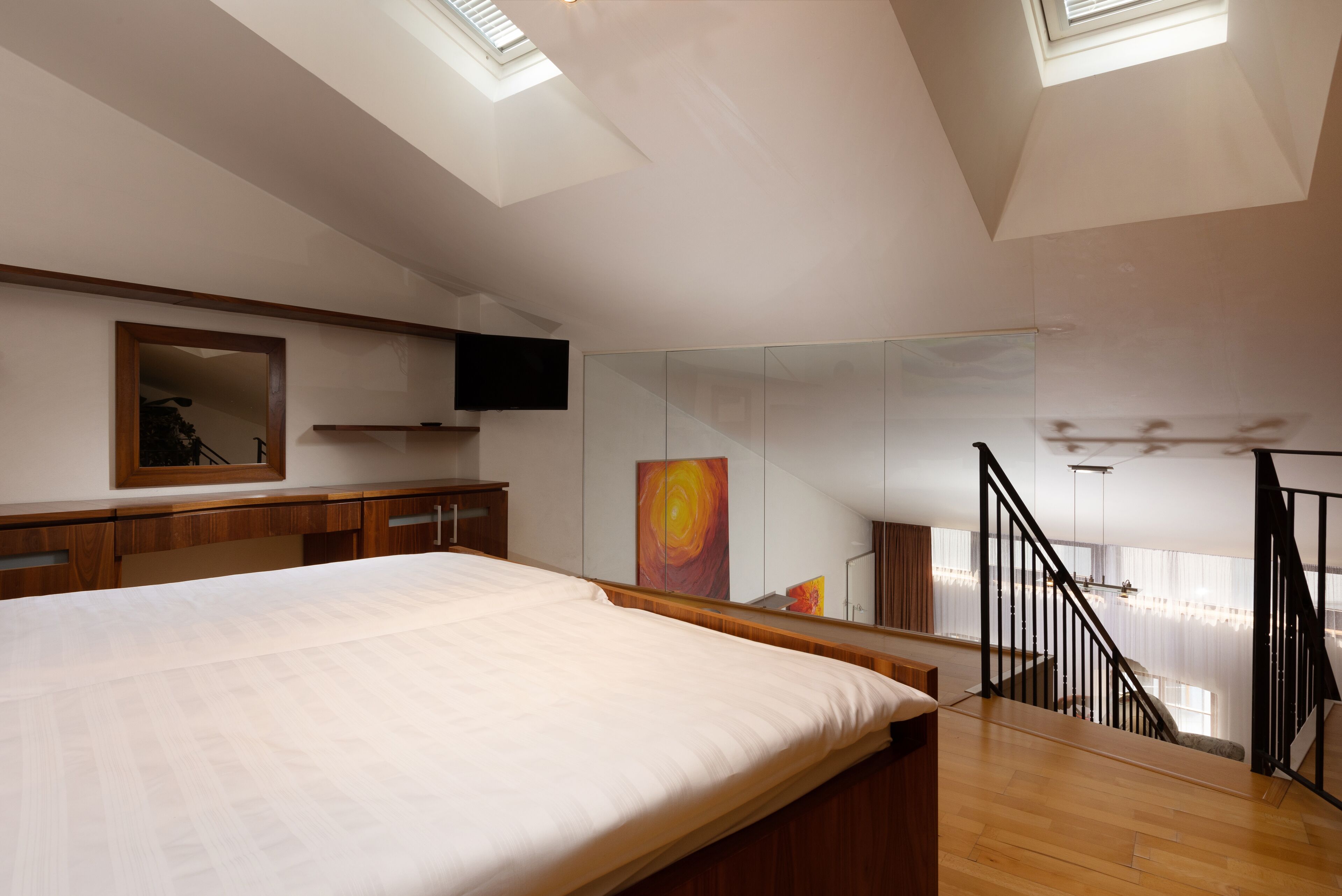 Ein Schlafzimmer mit einem großen Bett, einem Holztisch, einem an der Wand befestigten Fernseher, einem Gemälde und einer Treppe, die zu einer oberen Ebene führt.