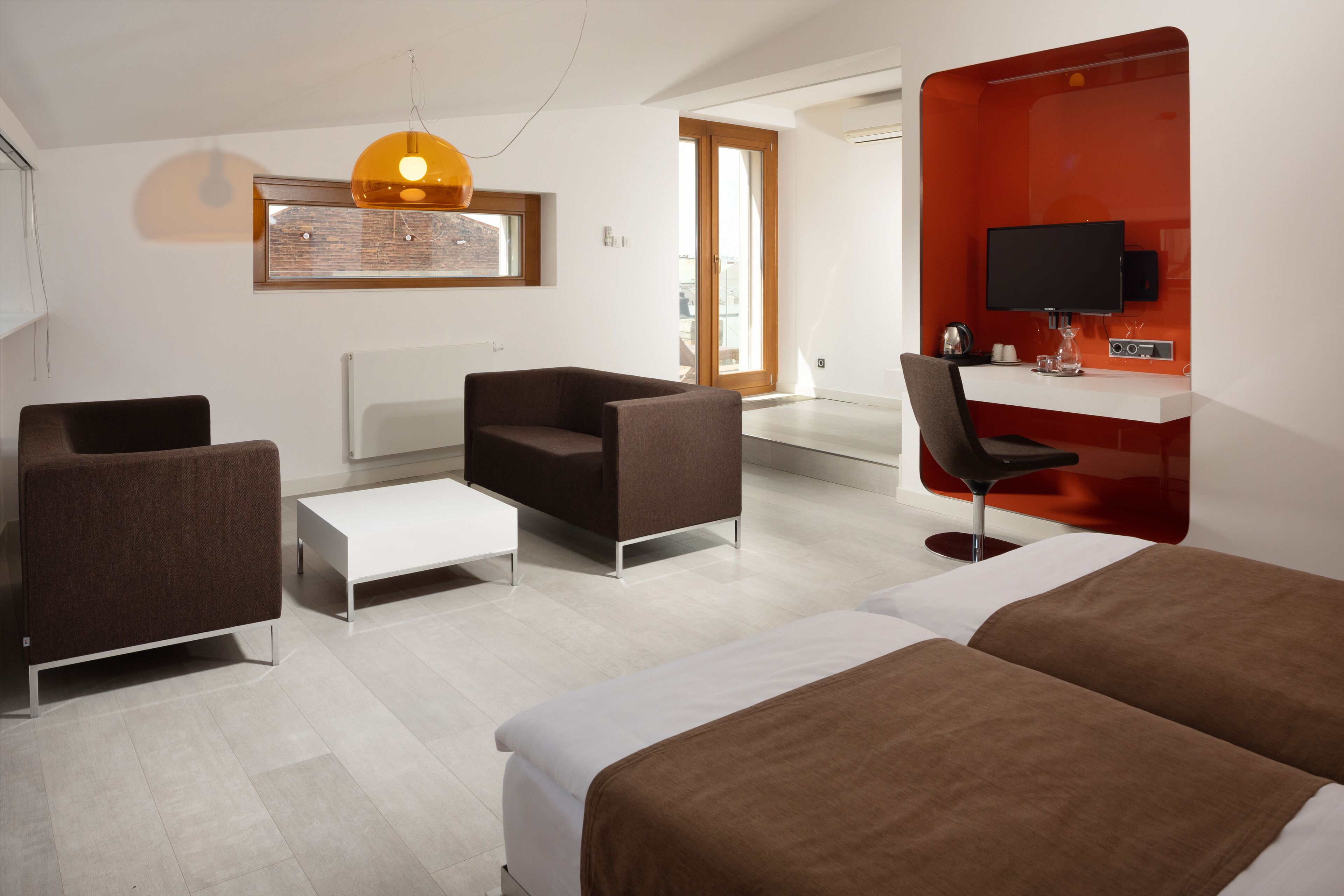 Quarto de hotel moderno com cama, duas poltronas, uma escrivaninha e uma televisão.