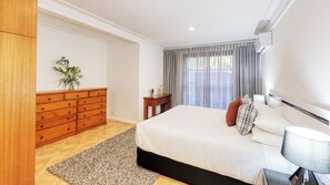 A bedroom with a bed, a wooden dresser, a nightstand, a lamp, and a window with curtains. - Mt Tamborine Stonehaven Manor (Tamborine Mountain)