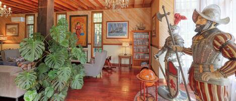 Una sala de estar acogedora con vigas de madera, un candelabro, una planta grande, una estantería y una estatua de un caballero.