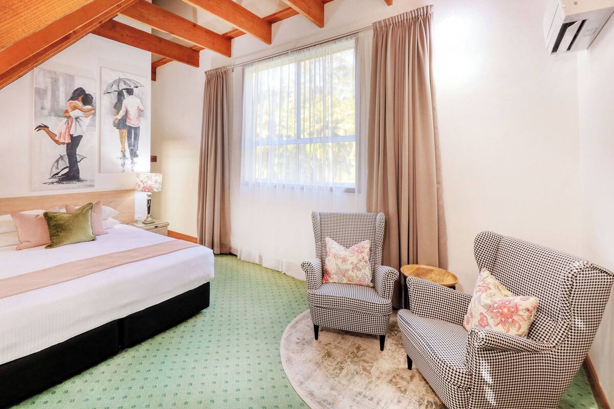 A hotel room with a large bed, two chairs, a small table, a window with curtains, and artwork on the wall.