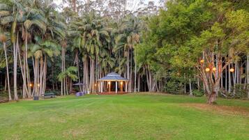 A gazebo with a roof and columns, surrounded by a lawn and tall trees.