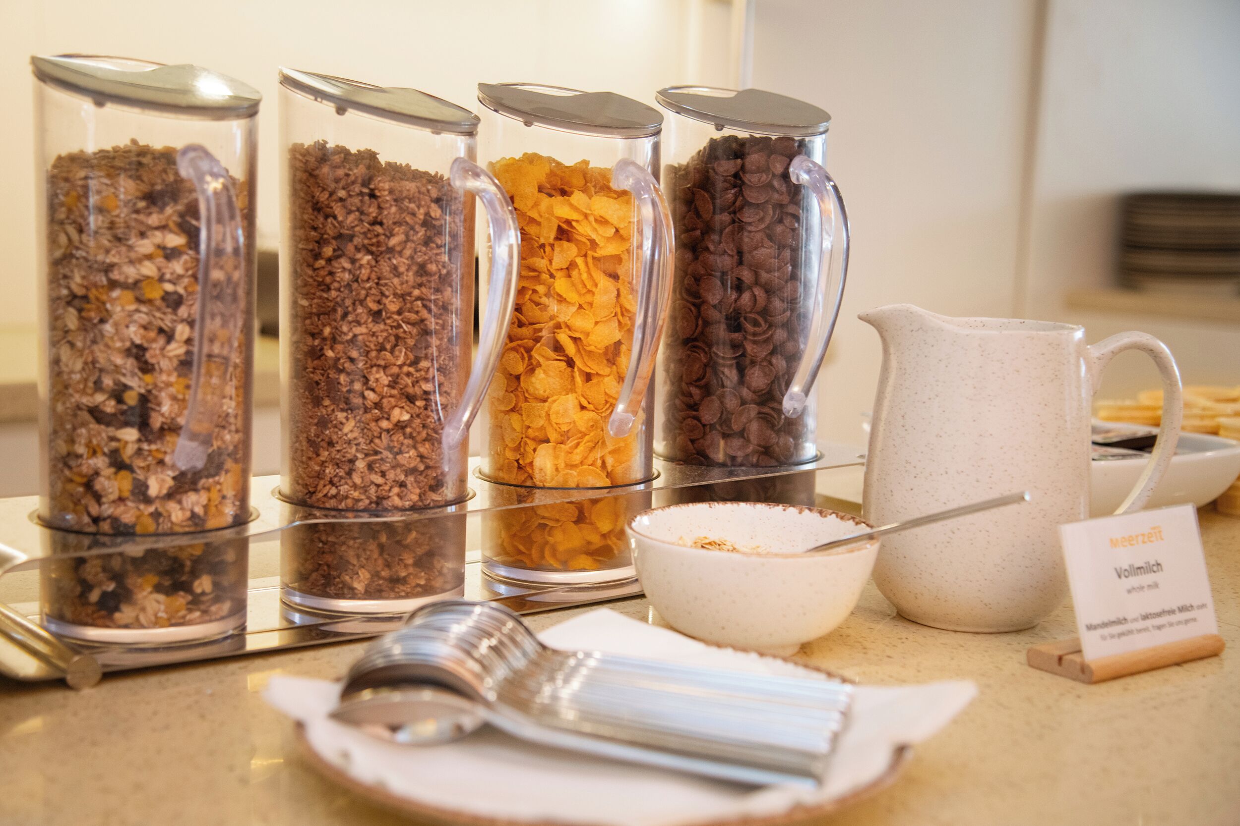 Un bufé de desayuno con diversas opciones de comida en recipientes transparentes, un tazón de cereales, una jarra de leche y un cartel que indica el tipo de leche.