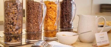 Un buffet de petit-déjeuner proposant divers aliments présentés dans des récipients transparents, un bol de céréales, une carafe de lait et un panneau indiquant le type de lait.