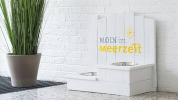 Una estación de alimentación para mascotas blanca con un cuenco metálico y un bebedero, con la etiqueta "MOIN im Meerzeit Hotel" e ícono de sol.