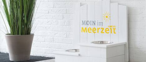 A white pet feeding station with a metal bowl and a water bowl, labeled "MOIN im Meerzeit Hotel" with a sun icon.