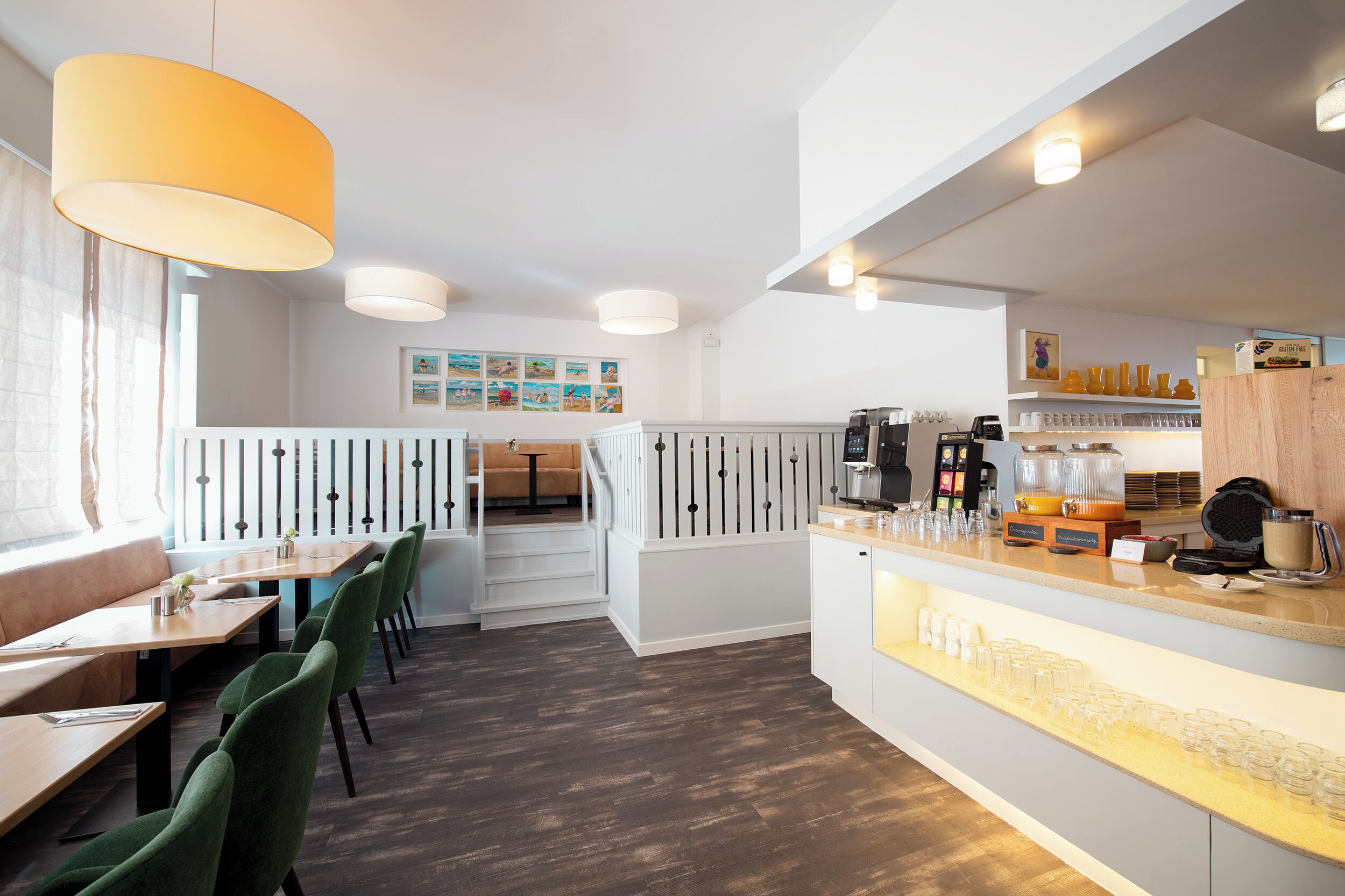 Un restaurante moderno con mesas de madera y sillas verdes, una barra con una máquina de café y una escalera que sube a un balcón.