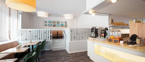 A modern restaurant with wooden tables and green chairs, a counter with a coffee machine, and a staircase leading to a balcony.