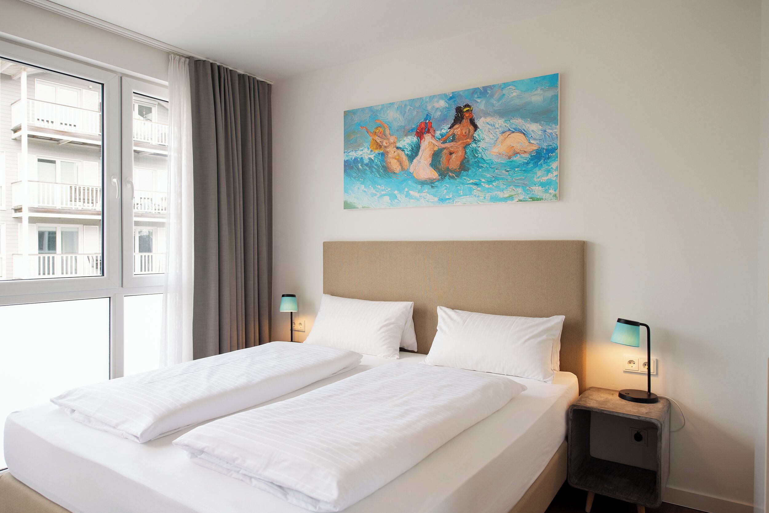 Habitación de hotel con una cama grande, dos mesitas de noche, un cuadro en la pared y una ventana con vistas a edificios.