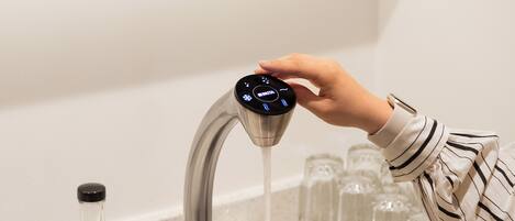 A hand adjusting a faucet with a digital interface, a glass carafe, and a row of glasses on a shelf.