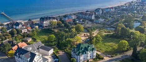 An aerial view of a coastal town with a mix of residential buildings, a large white building with a green roof, and a beach in the background.