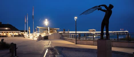 A waterfront promenade with a statue, a pier, and a building with outdoor seating.