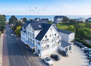 A two-story hotel with a balcony on the second floor, a parking lot with several cars, and a view of the sea in the background. - Hotel Meerzeit (Scharbeutz)