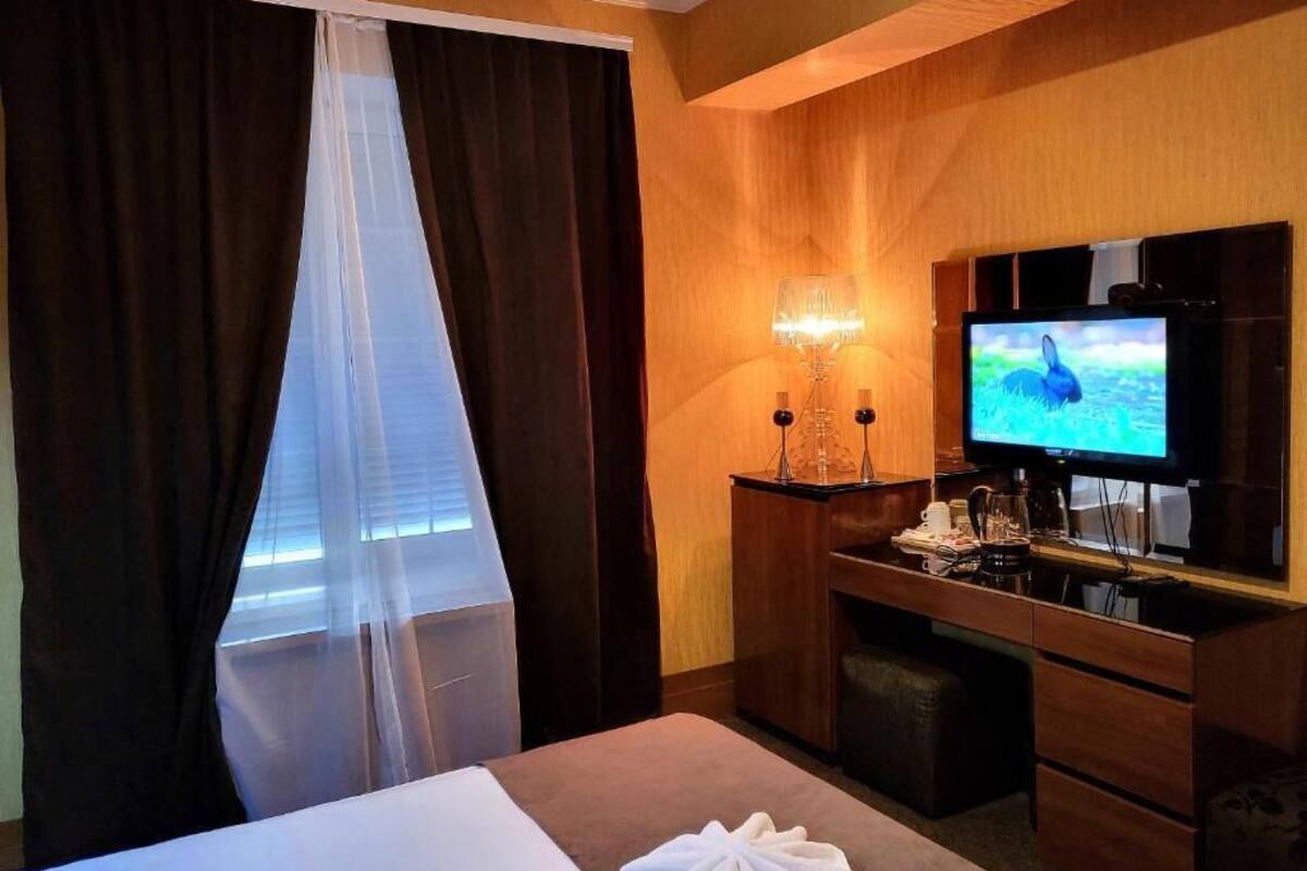 A hotel room with a bed, a TV, a desk with a lamp, and a window with curtains.