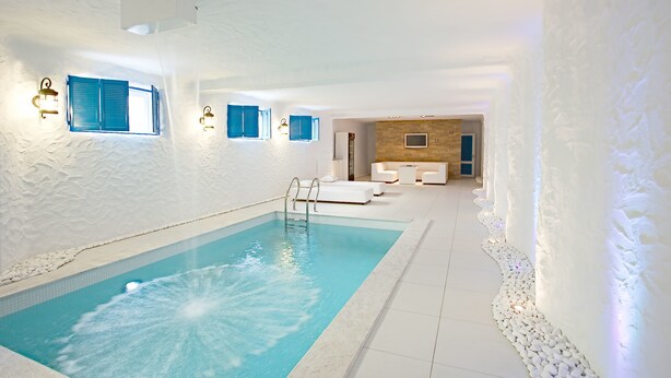 Piscina interior moderna com um recurso aquático de água azul cristalina, paredes brancas e área de estar com mobiliário branco.
