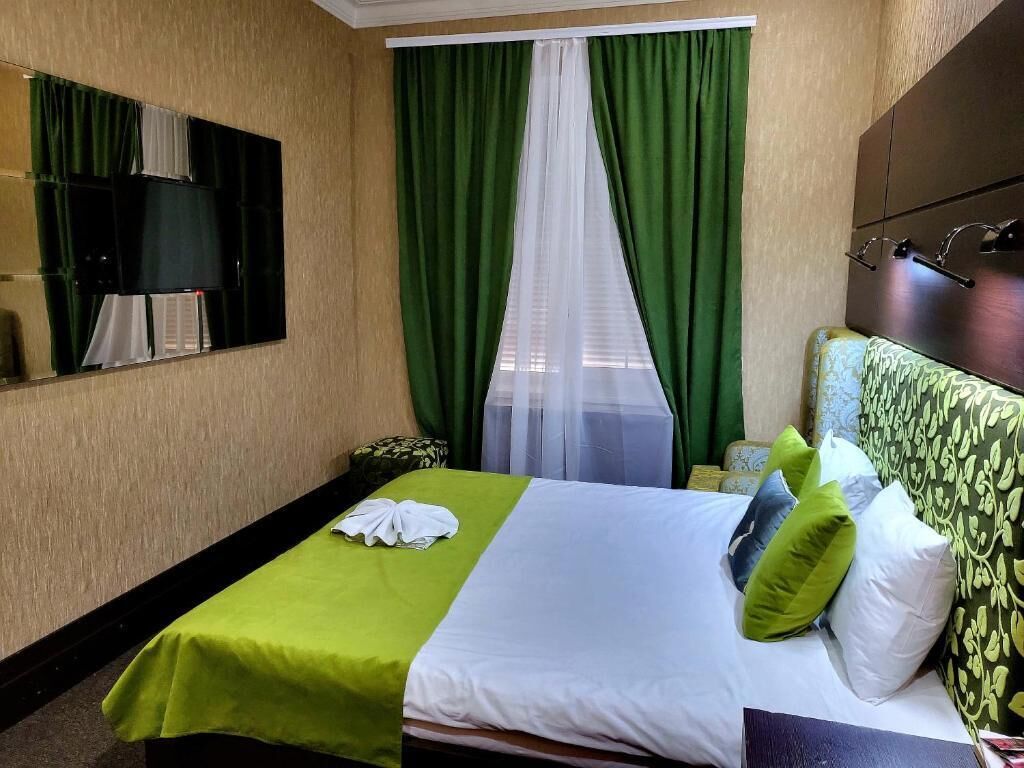 A hotel room with a bed, green curtains, a mosquito net, a bedside table, and a headboard with a patterned design.