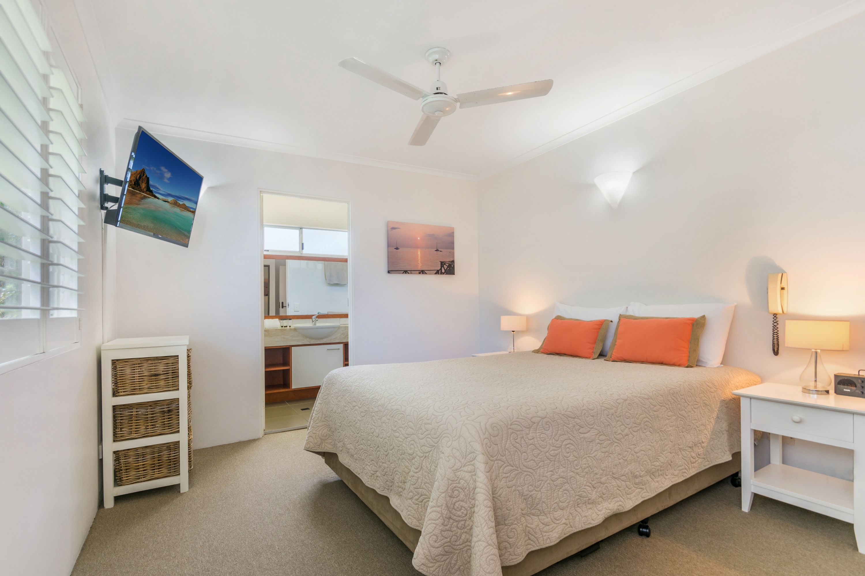 A bedroom with a bed, a nightstand, a ceiling fan, a wall-mounted TV, and a bathroom visible through an open door.