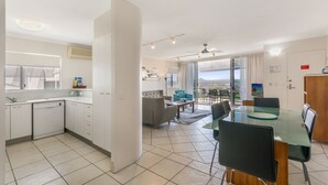 A modern kitchen with a dishwasher, a dining area with a glass table, and a view of the city. - Bali Hai Apartments Noosa (Noosa Heads)