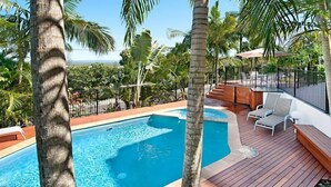 A swimming pool with a wooden deck, lounge chairs, and palm trees. - Bali Hai Apartments Noosa (Noosa Heads)