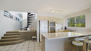 A modern kitchen with a central island, a built-in oven, and a refrigerator. There is a staircase leading to an upper level, and a window providing a view of greenery outside. - Bali Hai Apartments Noosa (Noosa Heads)