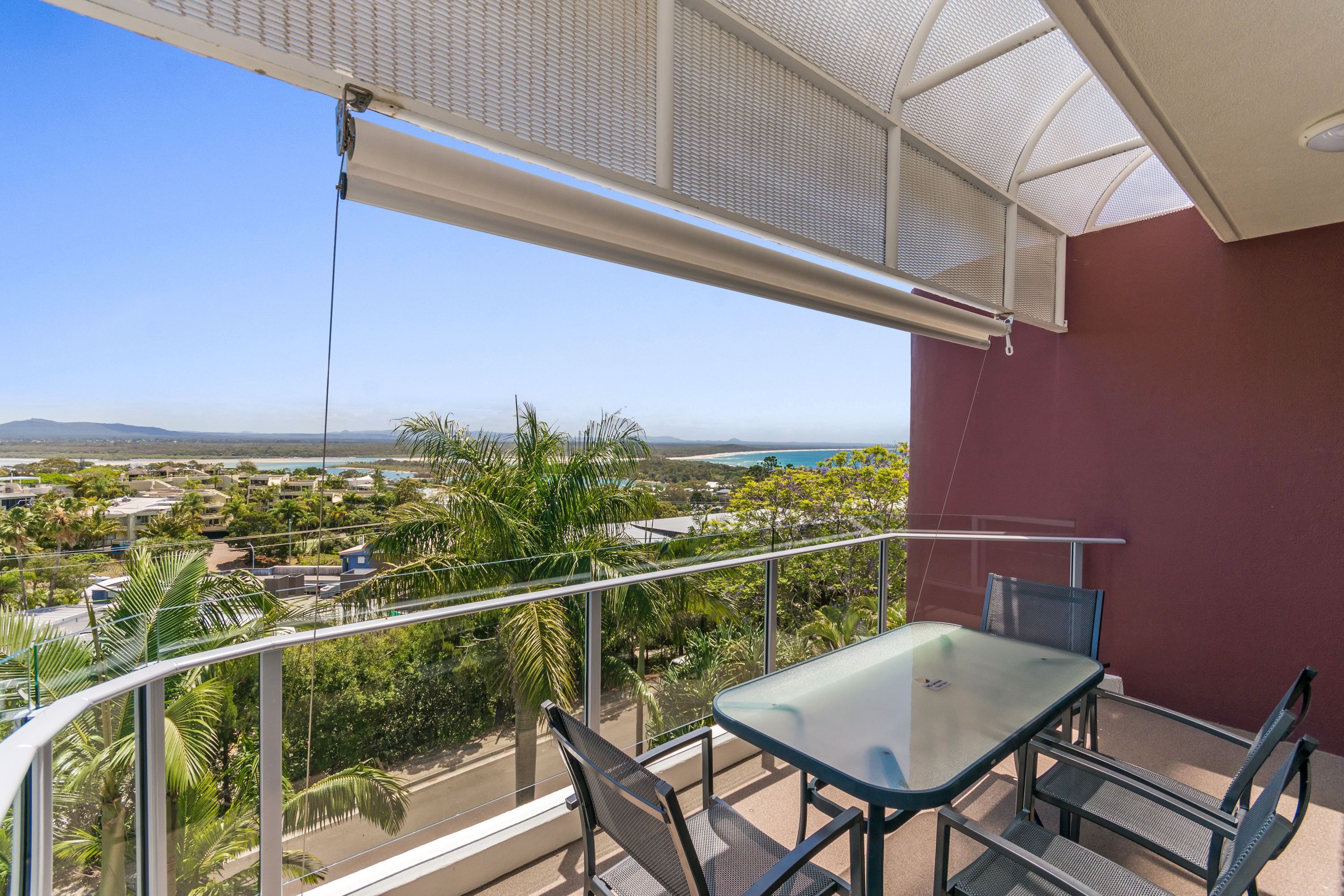 A balcony with a glass table and chairs, overlooking a coastal view with palm trees and buildings.