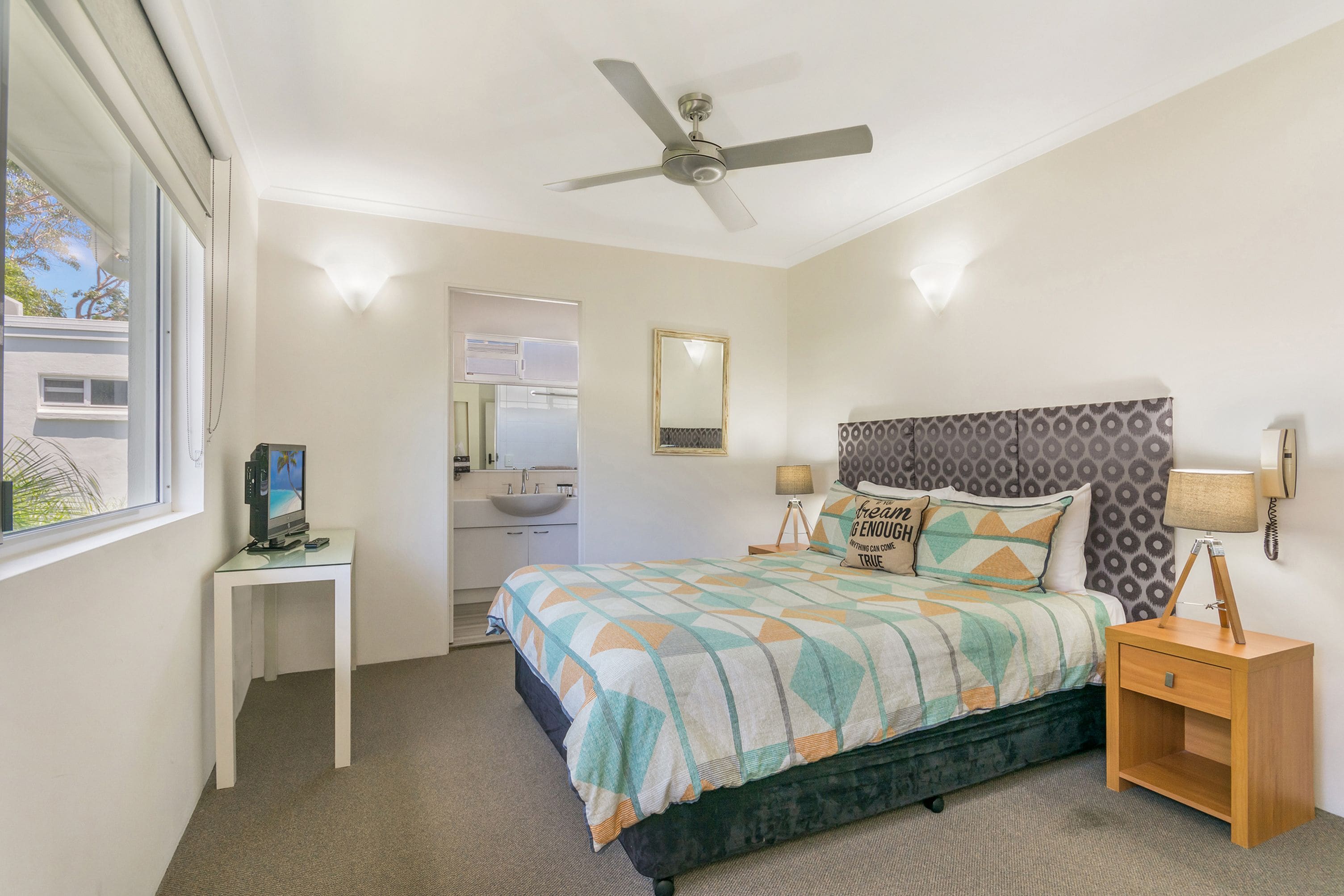 A bedroom with a bed, a desk, a nightstand, a ceiling fan, a mirror, and a window.