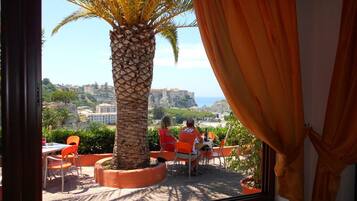 Una terraza con sillas color naranja y una palmera, con vistas panorámicas a un pueblo costero.