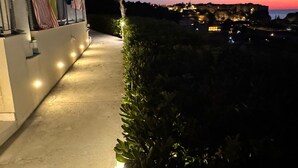 Vista nocturna de un edificio con balcón, un pequeño árbol y una ciudad iluminada a lo lejos.