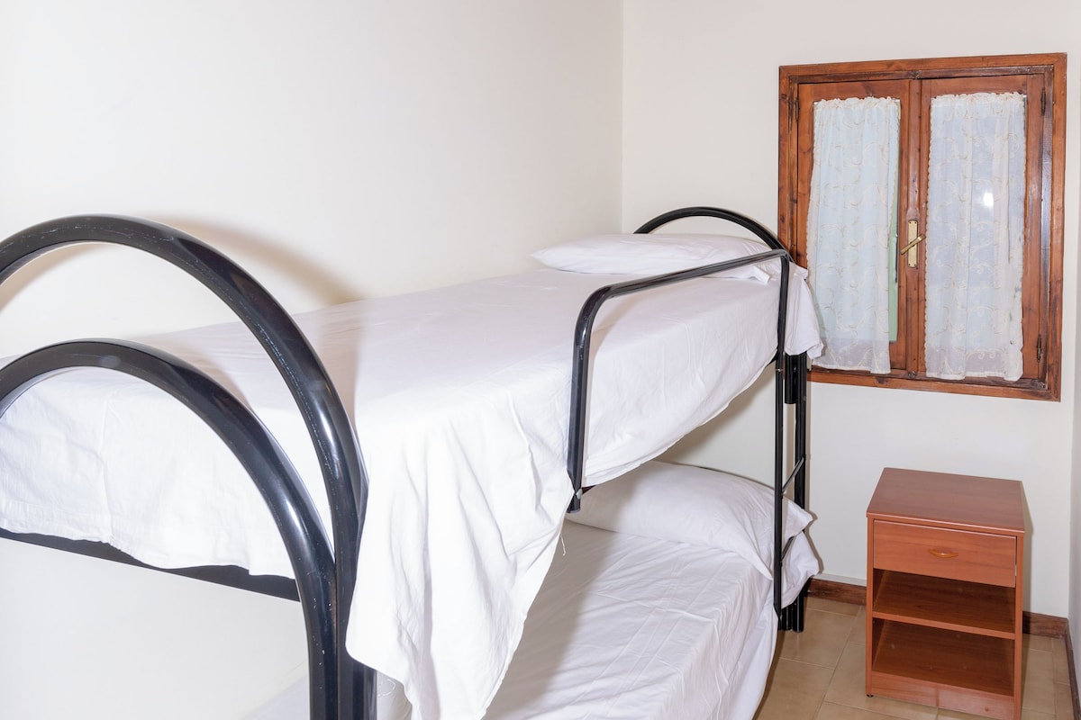 A bunk bed room with a wooden ceiling, a small wooden nightstand, and a window with curtains.