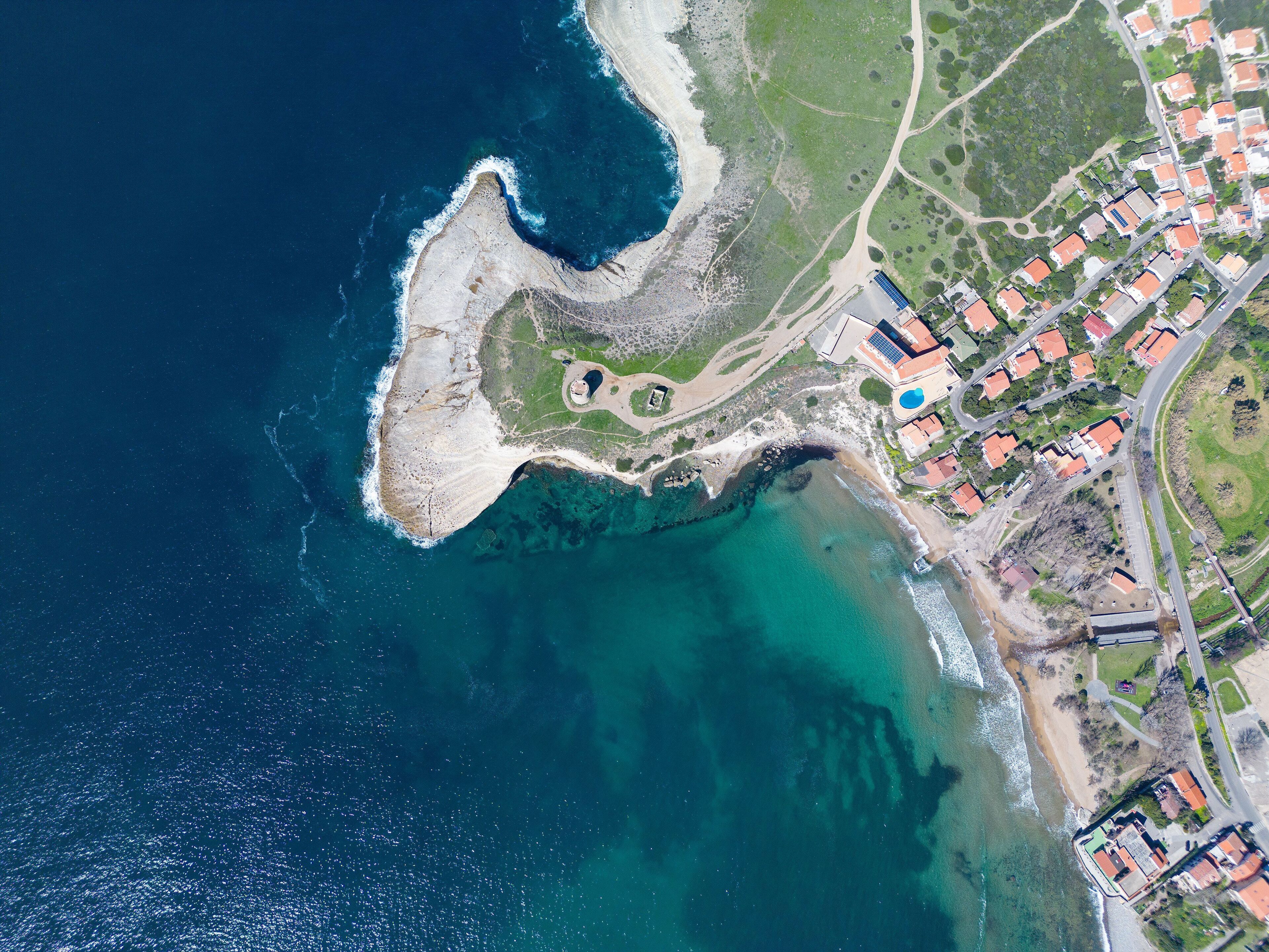 Une vue aérienne d’une zone côtière avec une plage, des eaux d’un bleu limpide et une zone résidentielle avec des maisons et une route.