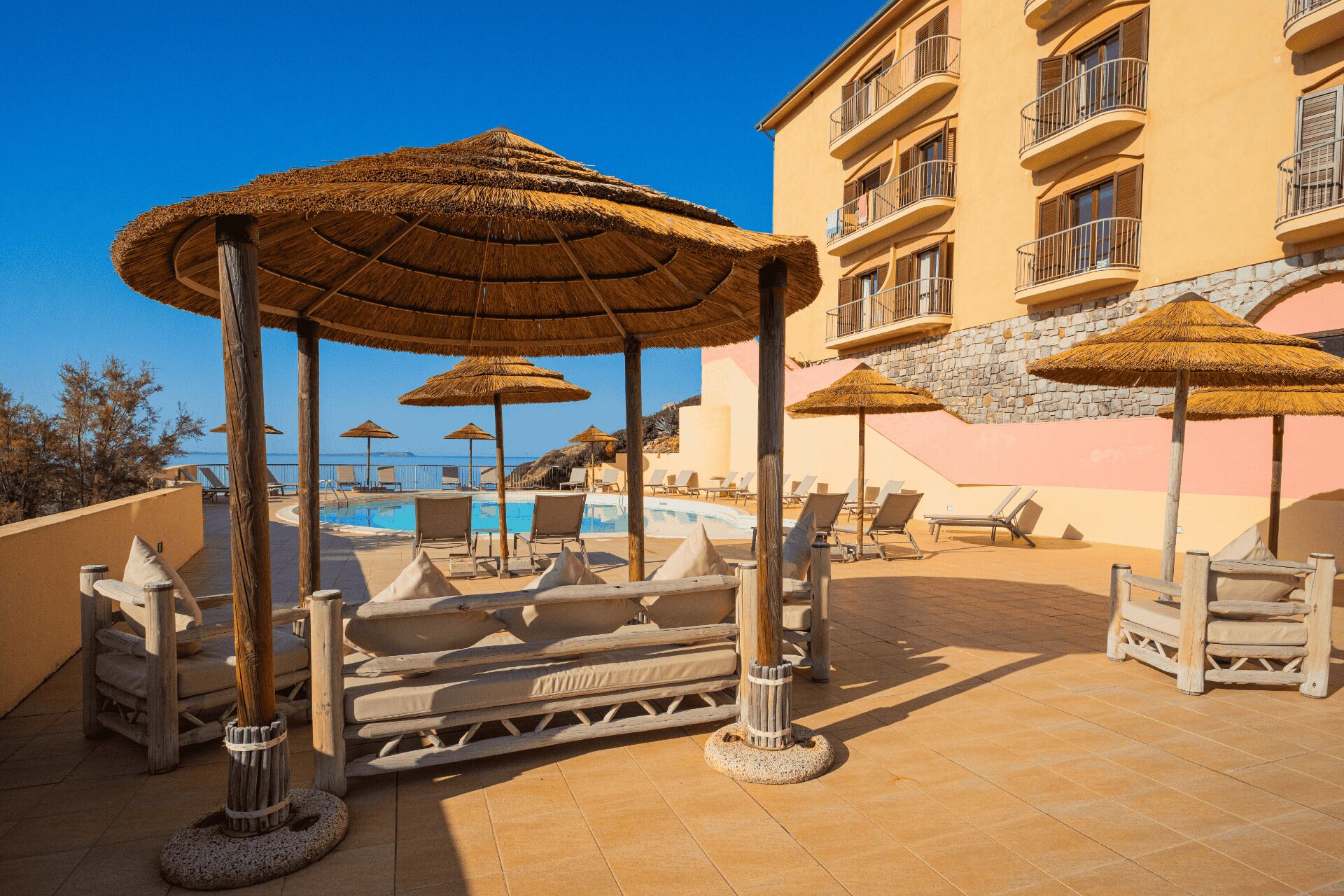 Un espace piscine aménagé dans un complexe hôtelier, avec des parasols en chaume, des chaises longues et un bâtiment doté de balcons.