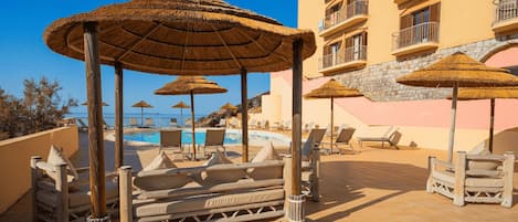 Un espace piscine aménagé dans un complexe hôtelier, avec des parasols en chaume, des chaises longues et un bâtiment doté de balcons.
