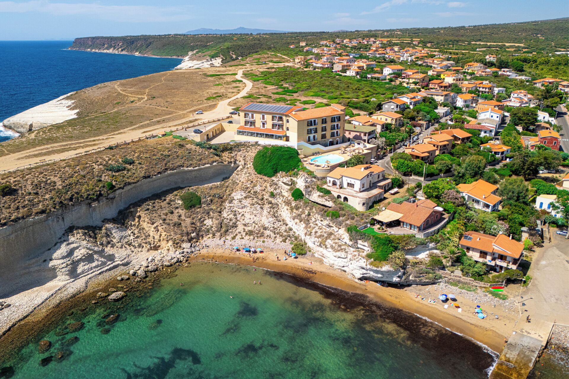 Luftaufnahme einer Küstenstadt mit Strand, Häusern an der Klippe und einem großen Gebäude mit Schwimmbad.