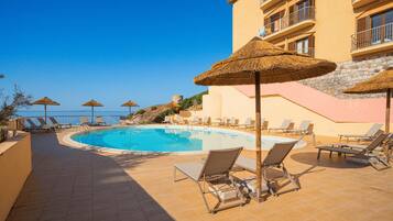 A pool area with lounge chairs and umbrellas, a clear view of the sea, and a building with balconies.