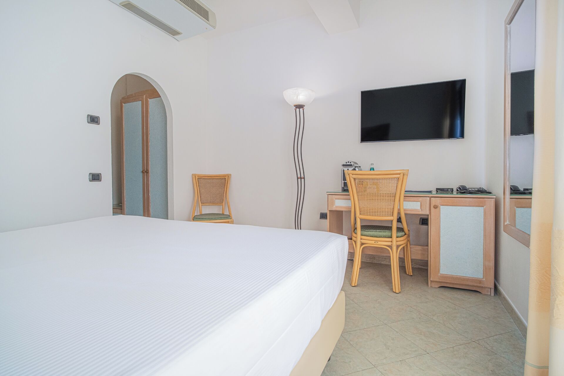 Habitación de hotel con una cama, dos sillones de mimbre, un escritorio con computadora, un televisor de pantalla plana y un espejo.