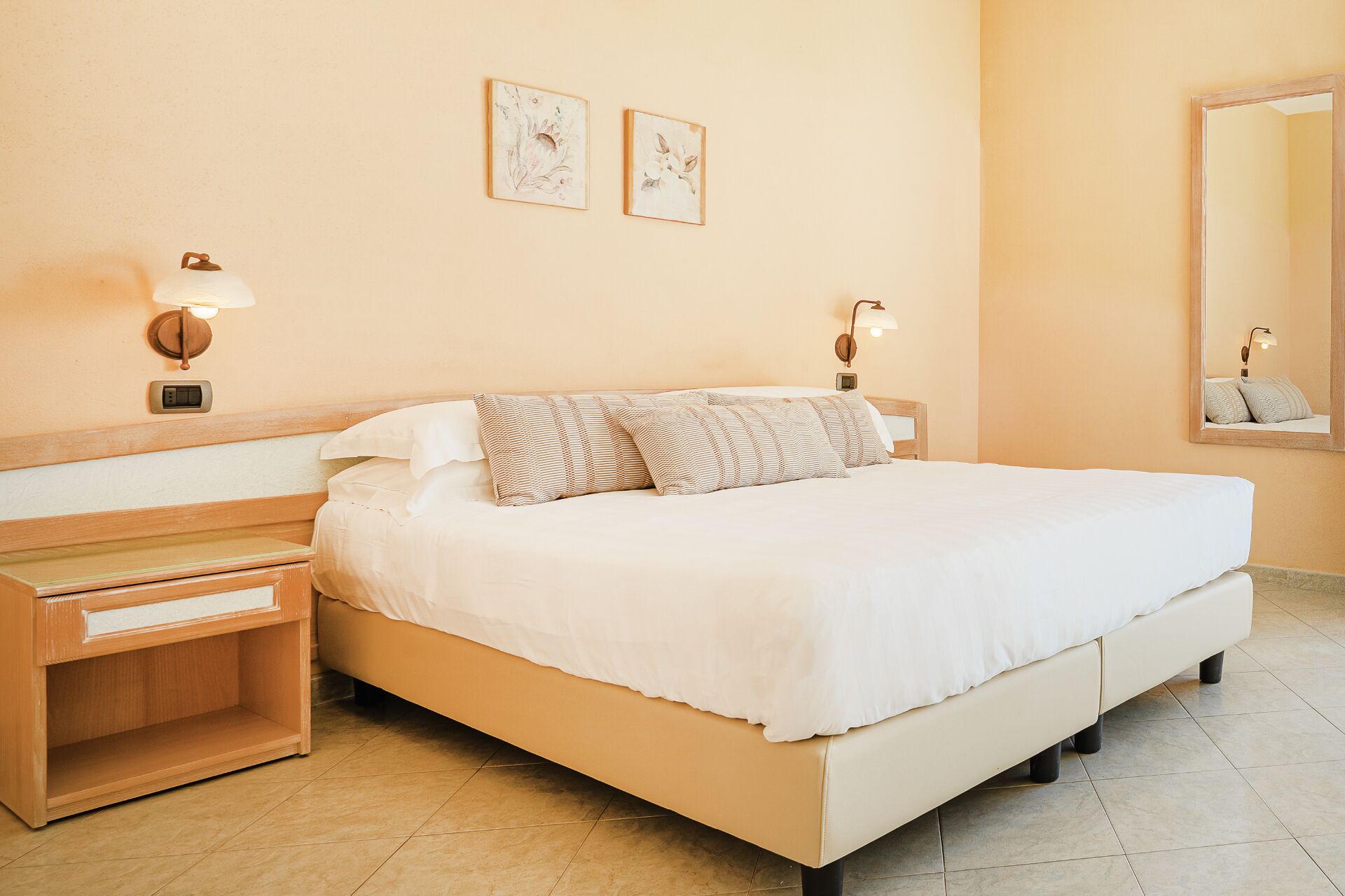 A hotel room with a large bed, bedside tables, wall-mounted lamps, and two framed floral artworks.