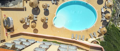 Vista aérea de un resort con una gran piscina rodeada de sillas de sol y sombrillas, un área de estar cubierta y una azotea con varios balcones.
