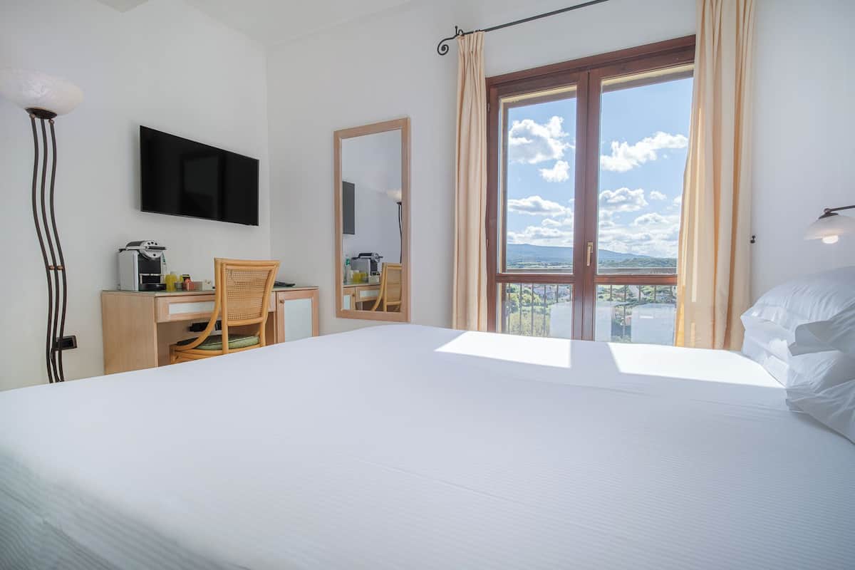 Ein Hotelzimmer mit Bett, Schreibtisch mit Stuhl, Flachbildfernseher, Spiegel und einem großen Fenster mit Blick auf Himmel und Wolken.