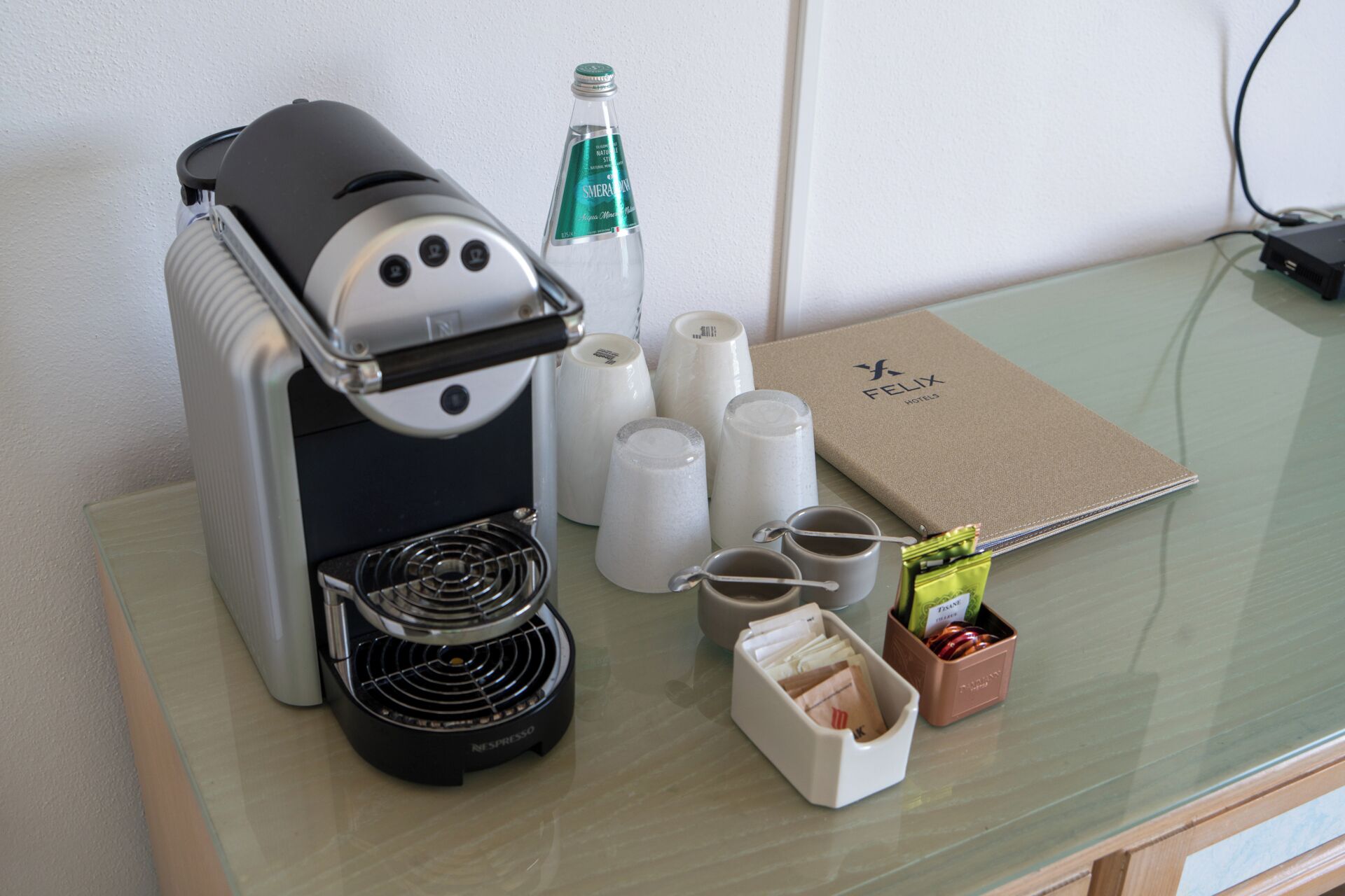 Una macchina per caffè Nespresso, una bottiglia d'acqua, una scatola di capsule per caffè, una capsula, una tazza da caffè, un cucchiaino e un kit di benvenuto per gli ospiti su un tavolo in vetro.