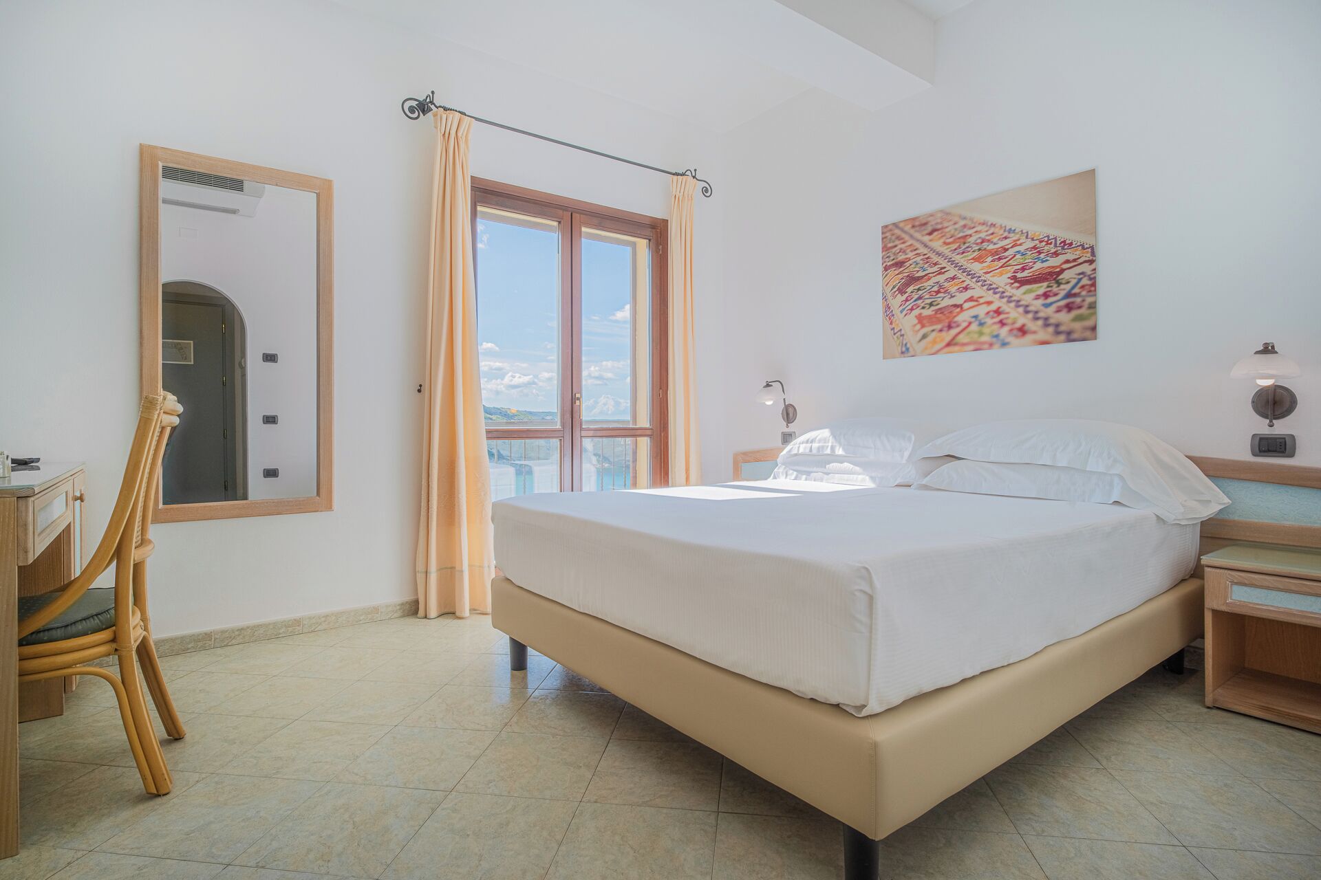 A bedroom with a bed, a desk, a chair, a window with curtains, a wall-mounted lamp, and a painting.