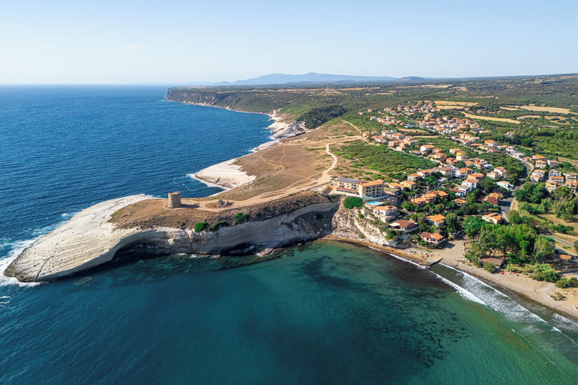 An aerial view of a coastal town with a prominent cliff, clear blue sea, and a densely built residential area with visible buildings and roads.